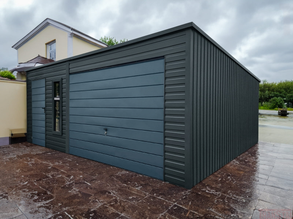 Modern matte black metal garage with flat roof, horizontal panel door, side access door, and vertical window, placed next to a cream house on a tiled driveway.